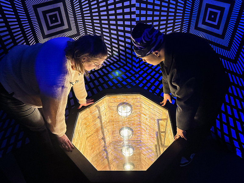 two women in a dark room with walls covered in geometric patterns staring down the tunnel of the ladder that journeys down into the centre of the earth