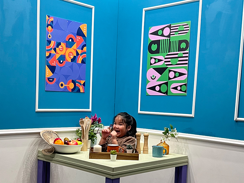 young girl with head popping through the centre of a dining room-style table adorned with food and utensils positioned in front of a light blue wall containing two framed images of colourful abstract geometrical patterns