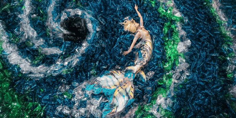 Overhead view of an art installation showing a mermaid‑like figure lying among swirling layers of blue, green, and clear plastic waste. The plastic is arranged to resemble ocean currents, with the figure’s flowing tail blending into the surrounding materials.