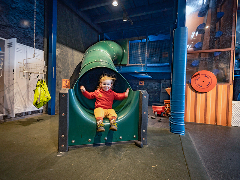 A young child in a red sweatshirt and yellow pants slides out of the mouth of a large green spiral tube slide inside an indoor play/exhibit area. The child’s arms are raised and they are smiling. The space around the slide has blue metal framing, padded safety posts, and a dark rubber floor; lockers and hanging high-visibility vests are visible on the left.