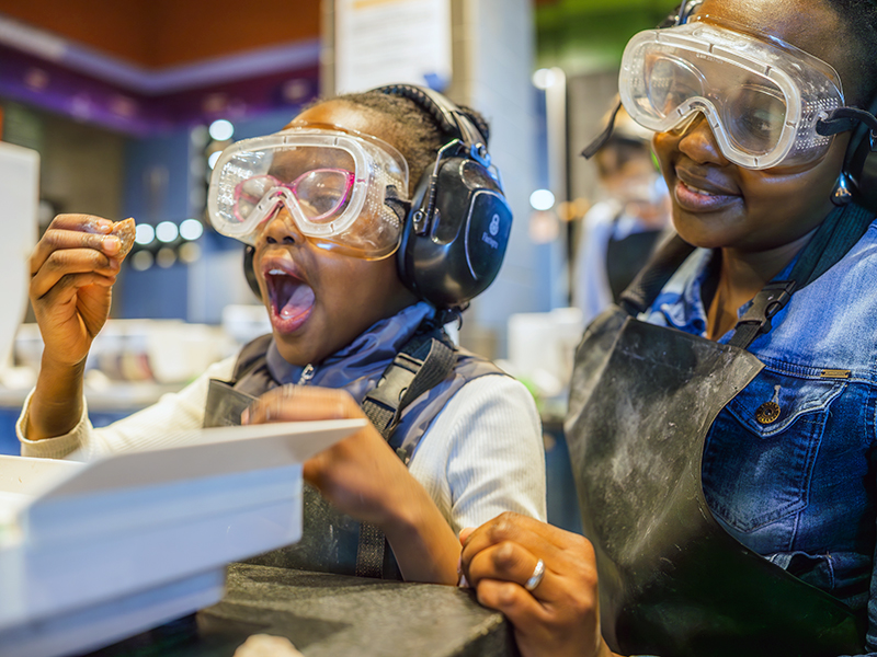 Close-up of two visitors at a hands-on exhibit wearing clear safety goggles, protective aprons, and earmuffs. The child in the foreground holds a small rock or mineral up for a closer look, mouth open in excitement or surprise. An adult beside them smiles while watching, with the exhibit workspace and blurred museum background behind them.