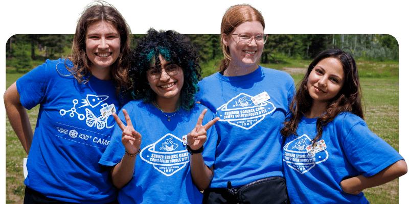 Four Summer Science Camp staff pose outdoors in blue camp shirts; one flashes a peace sign as they smile at the camera