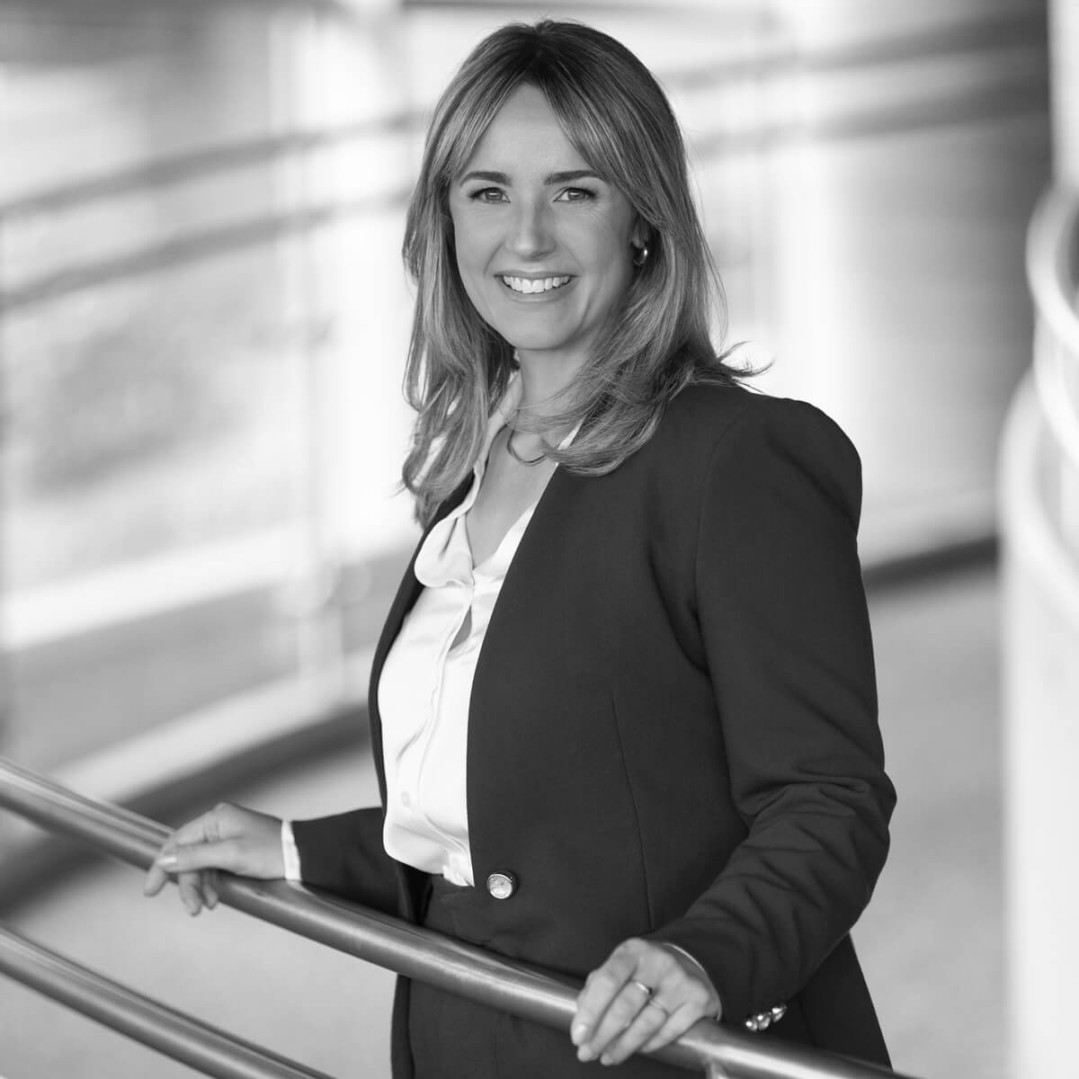 Professional portrait of Ashley Larose, CEO of Science North, smiling while standing on the Science North ramp. She is wearing a dark blazer over a light-coloured blouse, with the distinctive curved architecture of the science centre softly visible in the background.