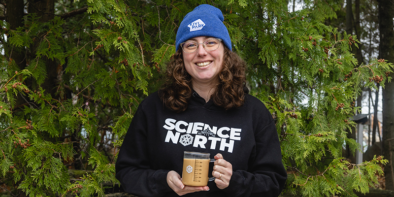 bluecoat in front of tree wearing science north toque and holding science north beaker mug